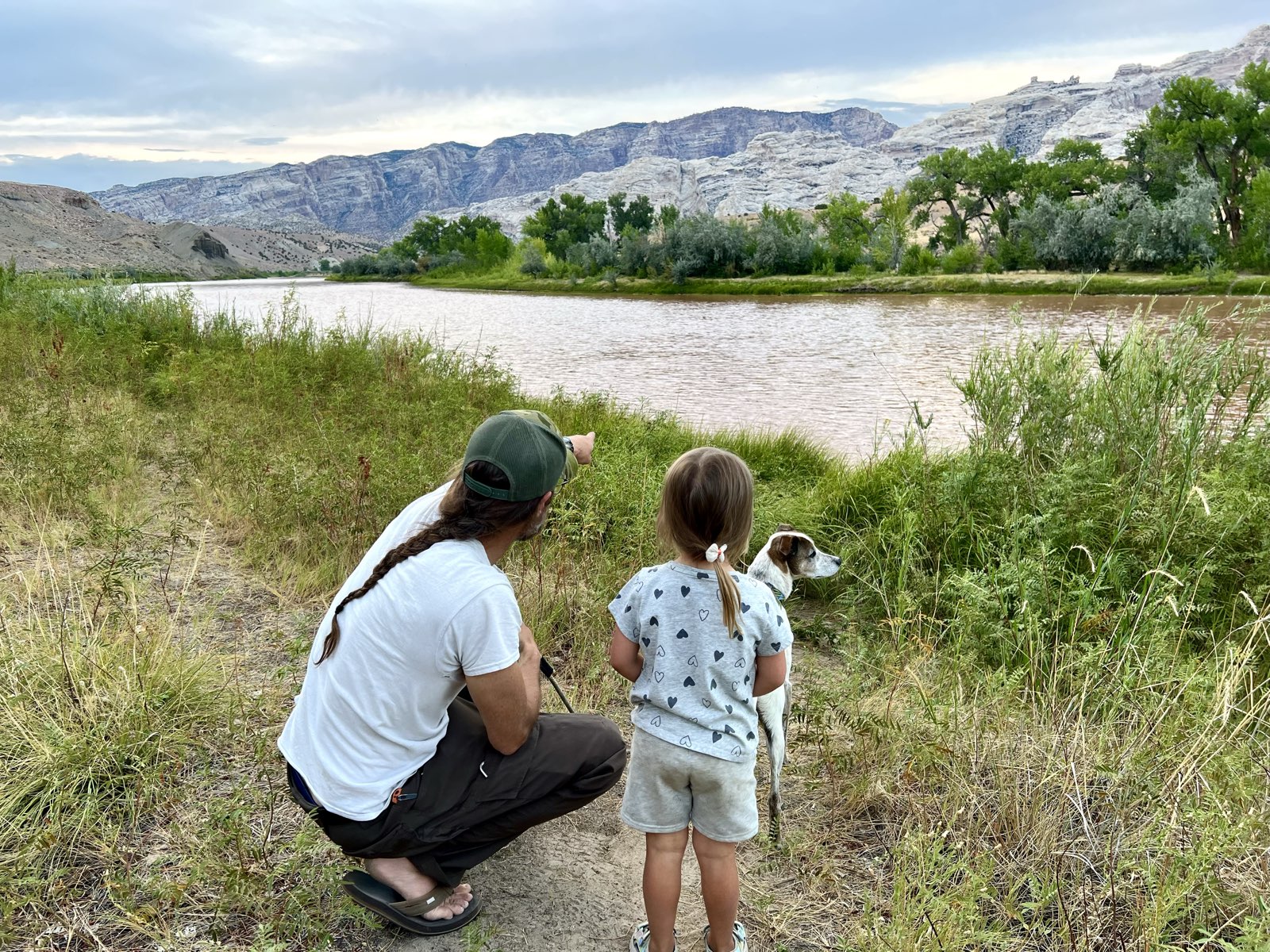 A man crouched on a riverbank beside a young girl and their dog, pointing across the water.
